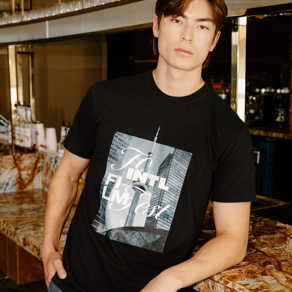 A man wearing a black graphic t-shirt with a New York City skyline print and gray striped pants, standing in a bar environment.