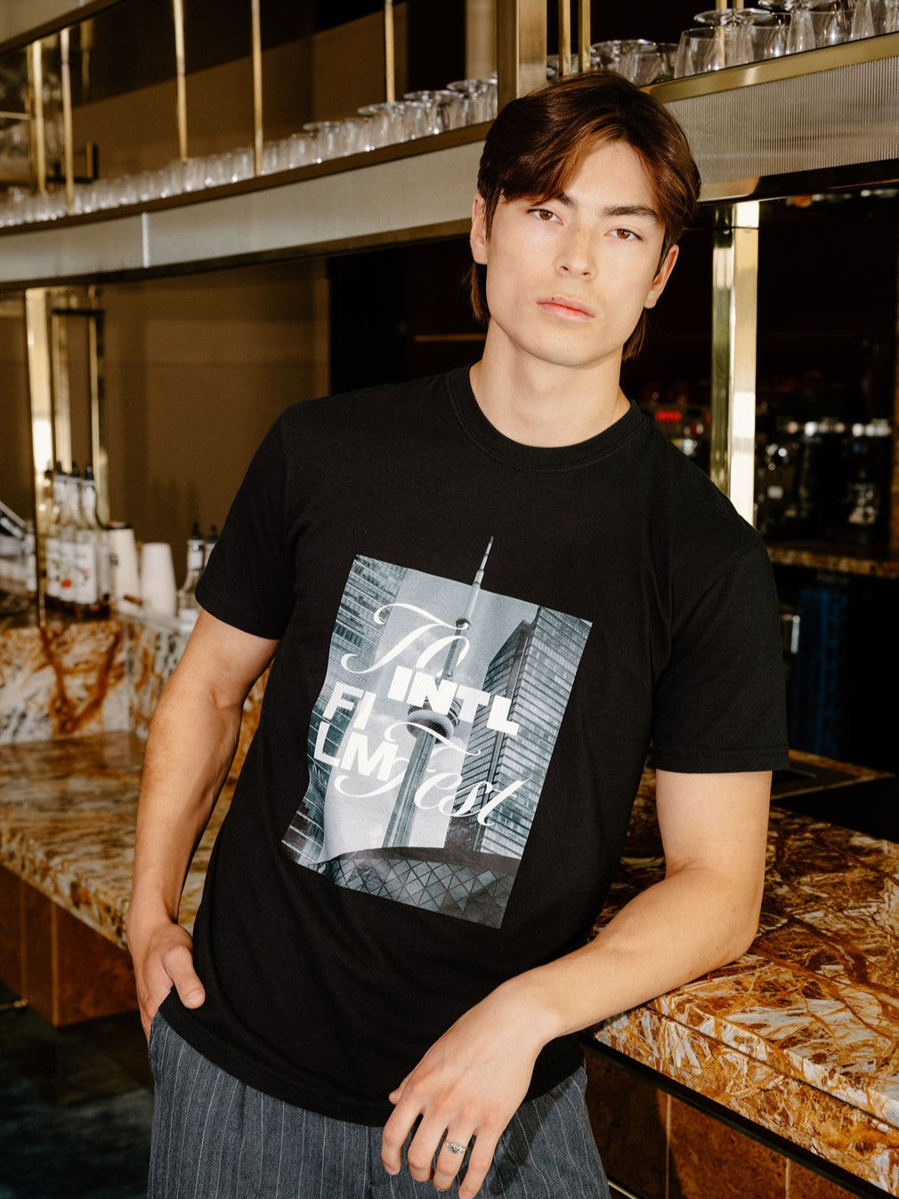 A man wearing a black graphic t-shirt with a New York City skyline print and gray striped pants, standing in a bar environment.