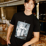 A man wearing a black graphic t-shirt with a New York City skyline print and gray striped pants, standing in a bar environment.