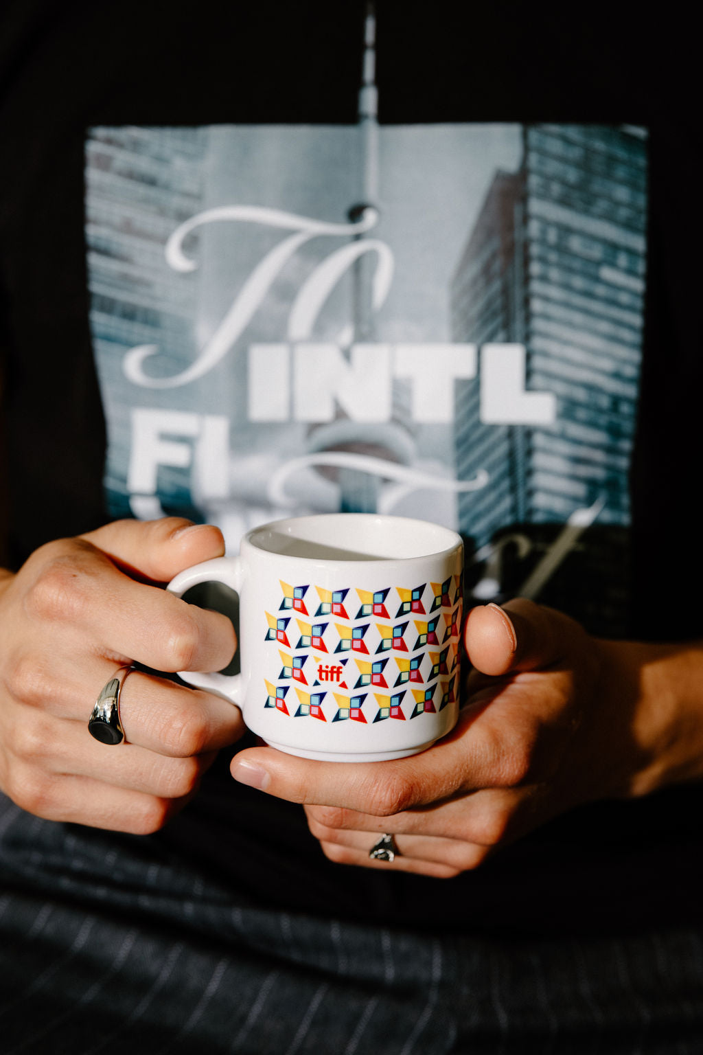 A person holding a white ceramic mug with a colorful geometric pattern of small flags. The person is wearing a black t-shirt with a graphic print.