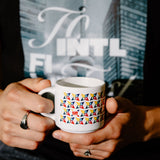 A person holding a white ceramic mug with a colorful geometric pattern of small flags. The person is wearing a black t-shirt with a graphic print.