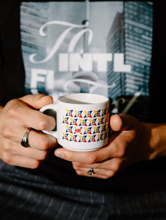 A person holding a white ceramic mug with a colorful geometric pattern of small flags. The person is wearing a black t-shirt with a graphic print.