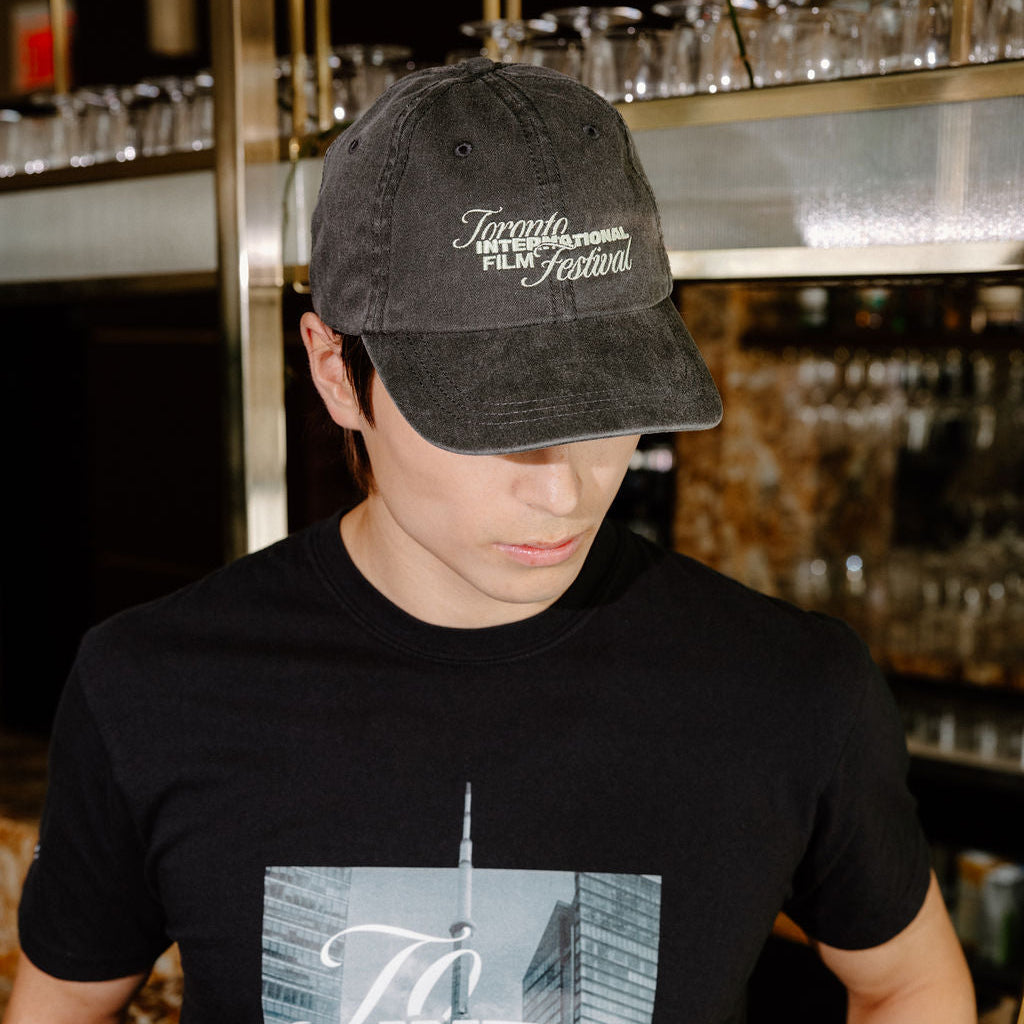 A man wearing a black t-shirt with a 'Toronto International Film Festival' graphic and a matching black baseball cap, standing in a bar environment.