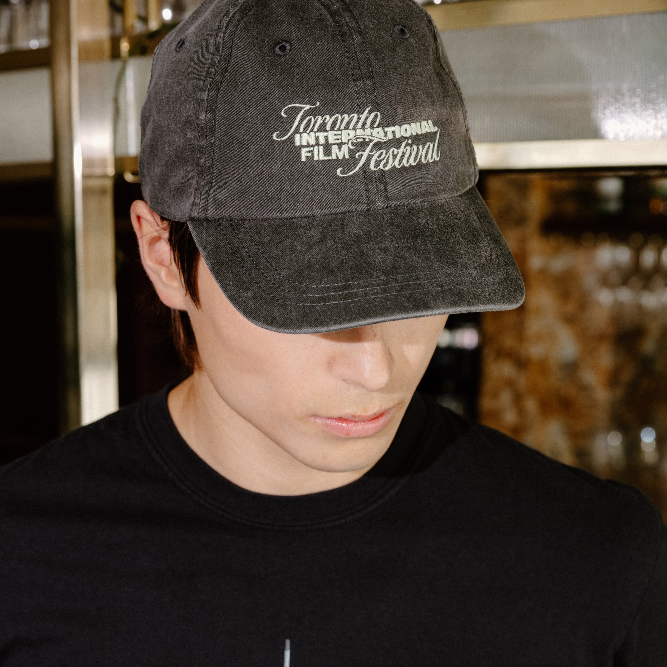 A man wearing a black t-shirt with a 'Toronto International Film Festival' graphic and a matching black baseball cap, standing in a bar environment.