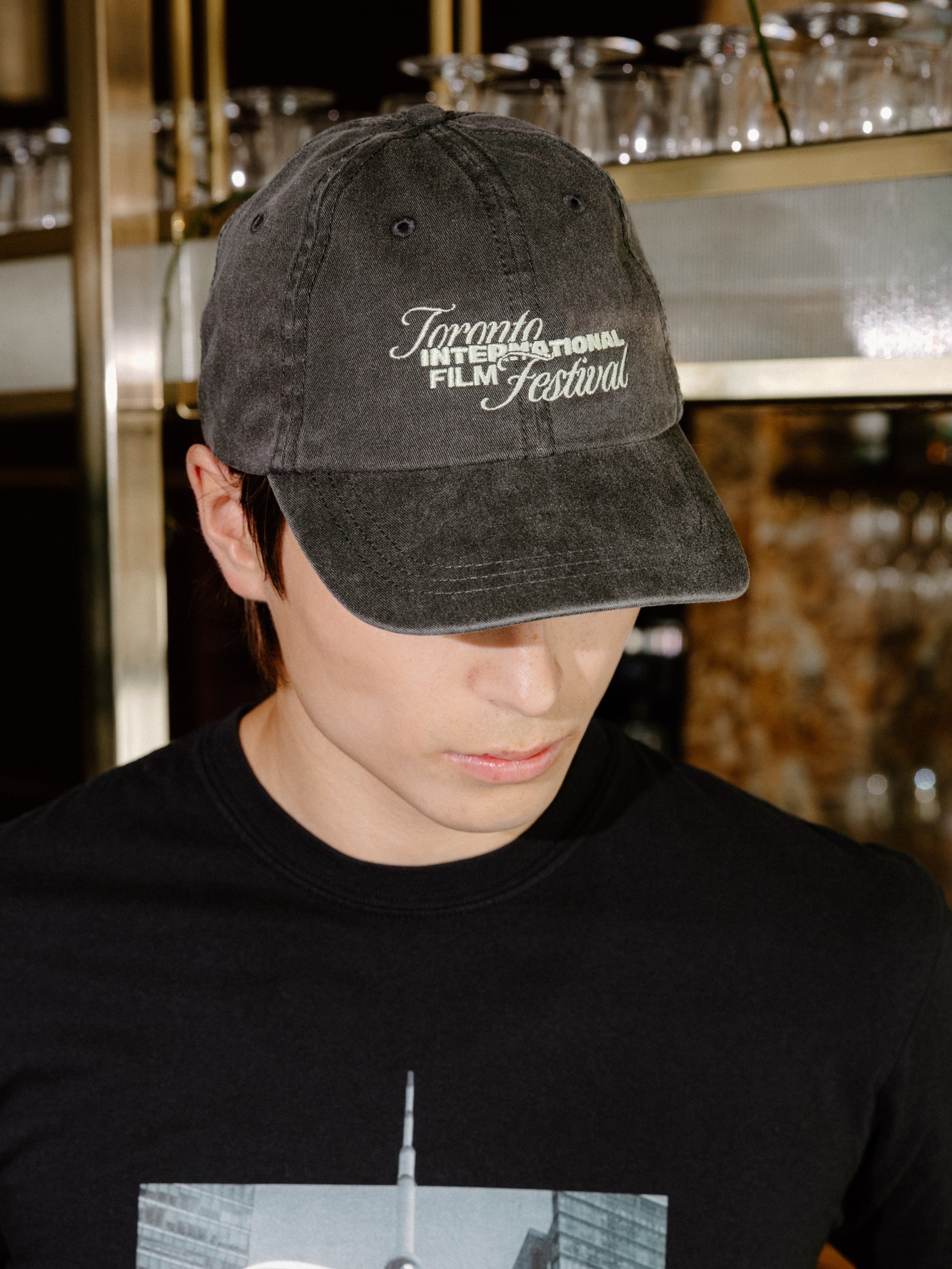 A man wearing a black t-shirt with a 'Toronto International Film Festival' graphic and a matching black baseball cap, standing in a bar environment.