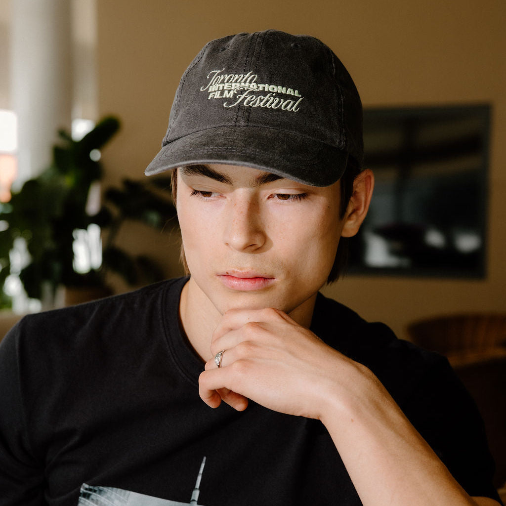A man wearing a black graphic t-shirt and a black baseball cap with 'Toronto International Film Festival' embroidered in white on the front, sitting indoors.