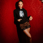 A woman posing in a dimly lit room with red patterned wallpaper, wearing a black graphic hoodie, brown lace-trimmed shorts, and black heels.