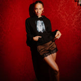 A woman posing in a dimly lit room with red patterned wallpaper, wearing a black graphic hoodie, brown lace-trimmed shorts, and black heels.