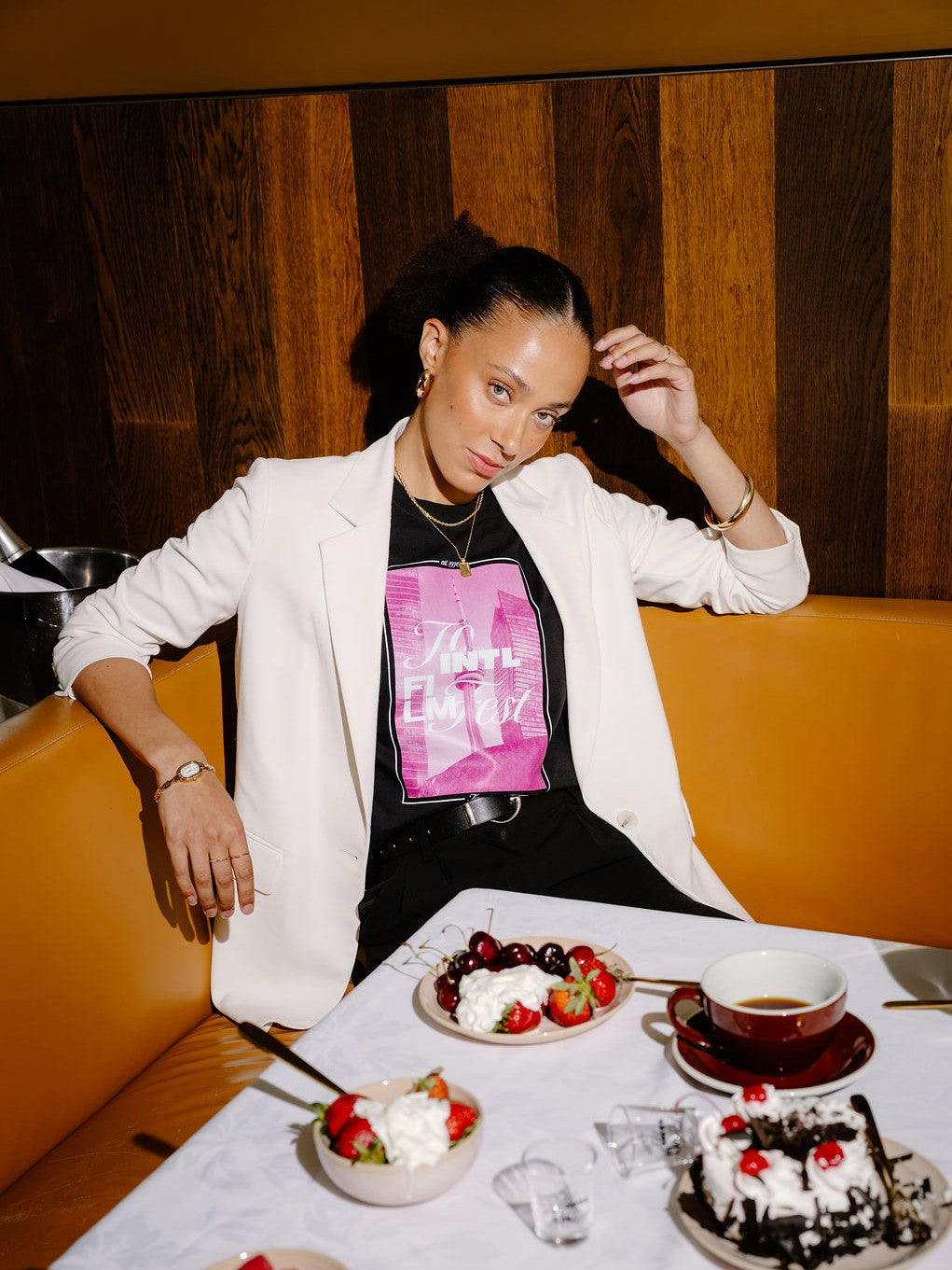 A woman posing in a restaurant booth wearing a white blazer over a black t-shirt with a pink graphic print. In front of her is a table set with desserts, including a chocolate cake with cherries, bowls of strawberries and whipped cream, and a cup of tea