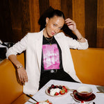A woman posing in a restaurant booth wearing a white blazer over a black t-shirt with a pink graphic print. In front of her is a table set with desserts, including a chocolate cake with cherries, bowls of strawberries and whipped cream, and a cup of tea