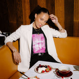 A woman posing in a restaurant booth wearing a white blazer over a black t-shirt with a pink graphic print. In front of her is a table set with desserts, including a chocolate cake with cherries, bowls of strawberries and whipped cream, and a cup of tea