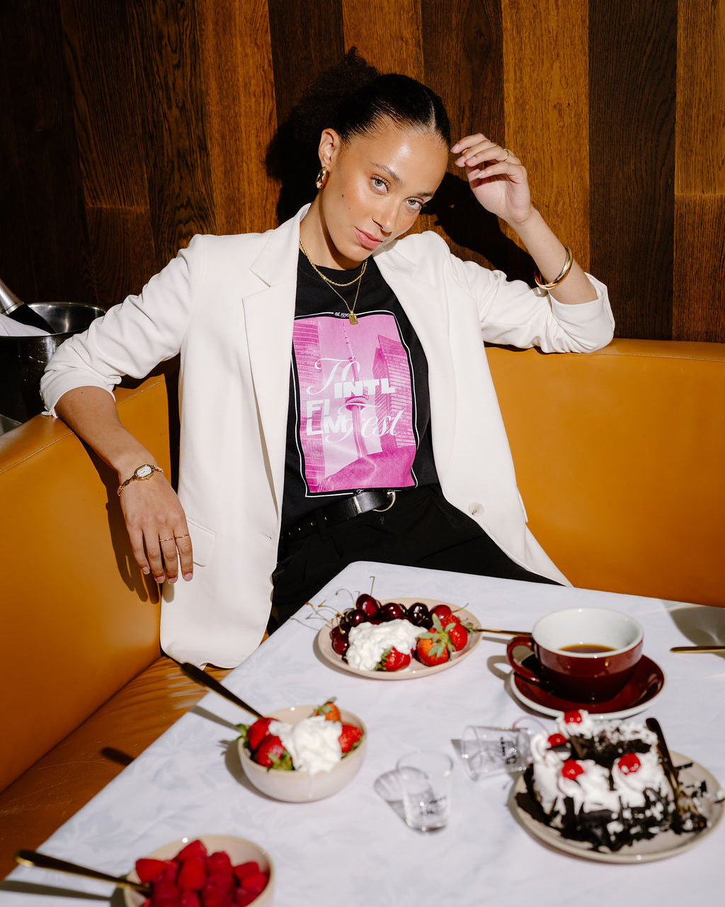 A woman posing in a restaurant booth wearing a white blazer over a black t-shirt with a pink graphic print. In front of her is a table set with desserts, including a chocolate cake with cherries, bowls of strawberries and whipped cream, and a cup of tea