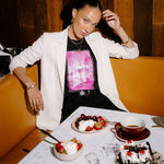 A woman posing in a restaurant booth wearing a white blazer over a black t-shirt with a pink graphic print. In front of her is a table set with desserts, including a chocolate cake with cherries, bowls of strawberries and whipped cream, and a cup of tea