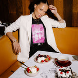 A woman posing in a restaurant booth wearing a white blazer over a black t-shirt with a pink graphic print. In front of her is a table set with desserts, including a chocolate cake with cherries, bowls of strawberries and whipped cream, and a cup of tea