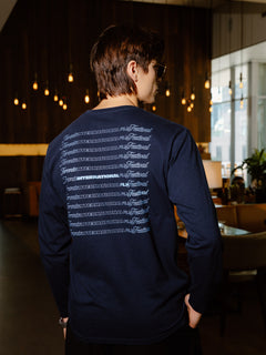 A man wearing a navy blue long-sleeve shirt with a white graphic print on the back, photographed from behind in a dimly lit indoor environment.
