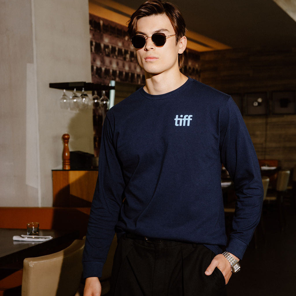 A man wearing a navy blue long-sleeve crewneck sweatshirt with a white 'tiff' logo on the chest, paired with black pants and sunglasses, standing in a dimly lit restaurant or bar environment.