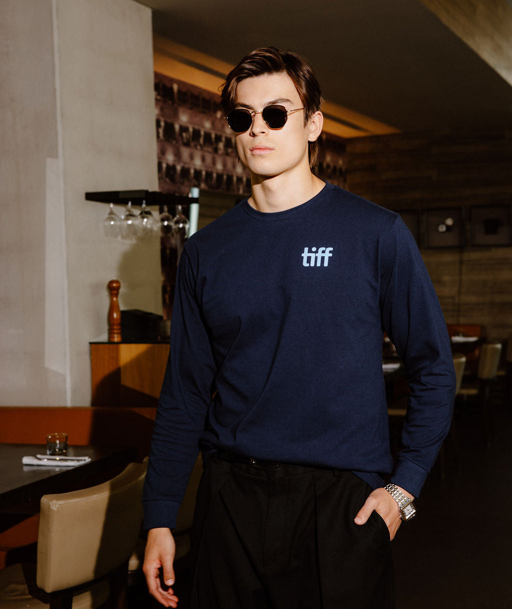A man wearing a navy blue long-sleeve crewneck sweatshirt with a white 'tiff' logo on the chest, paired with black pants and sunglasses, standing in a dimly lit restaurant or bar environment.