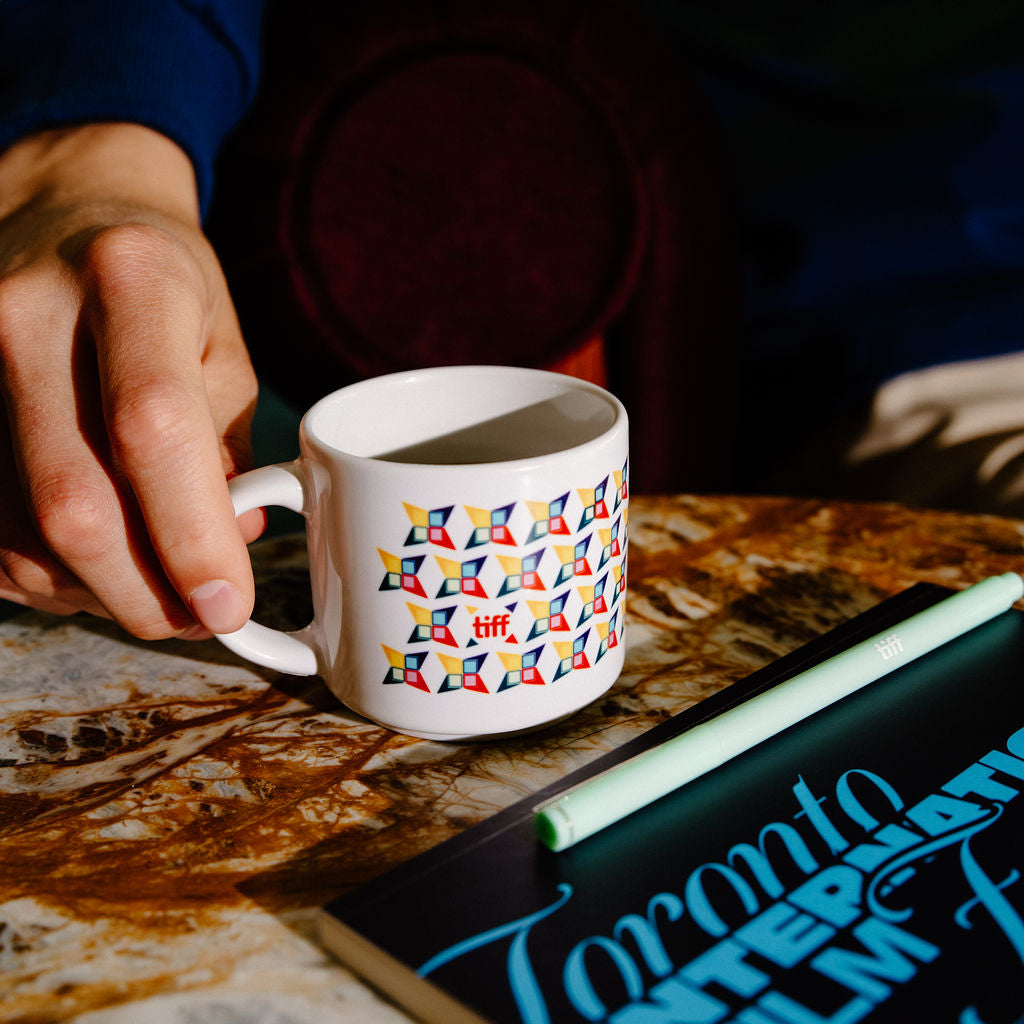 A white ceramic mug with a colorful geometric pattern, held by a hand over a marble table next to a notebook.