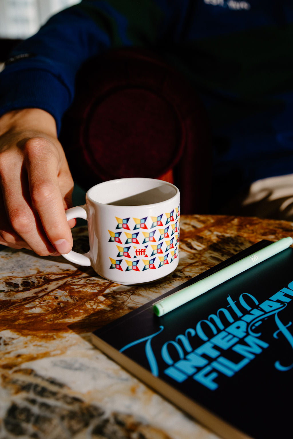 A white ceramic mug with a colorful geometric pattern, held by a hand over a marble table next to a notebook.