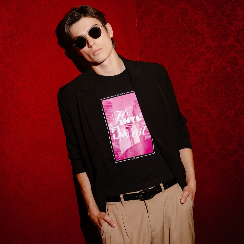 A man wearing a black long-sleeve blazer over a black graphic t-shirt with a pink and white print, paired with beige trousers and sunglasses, posing against a textured red wall.