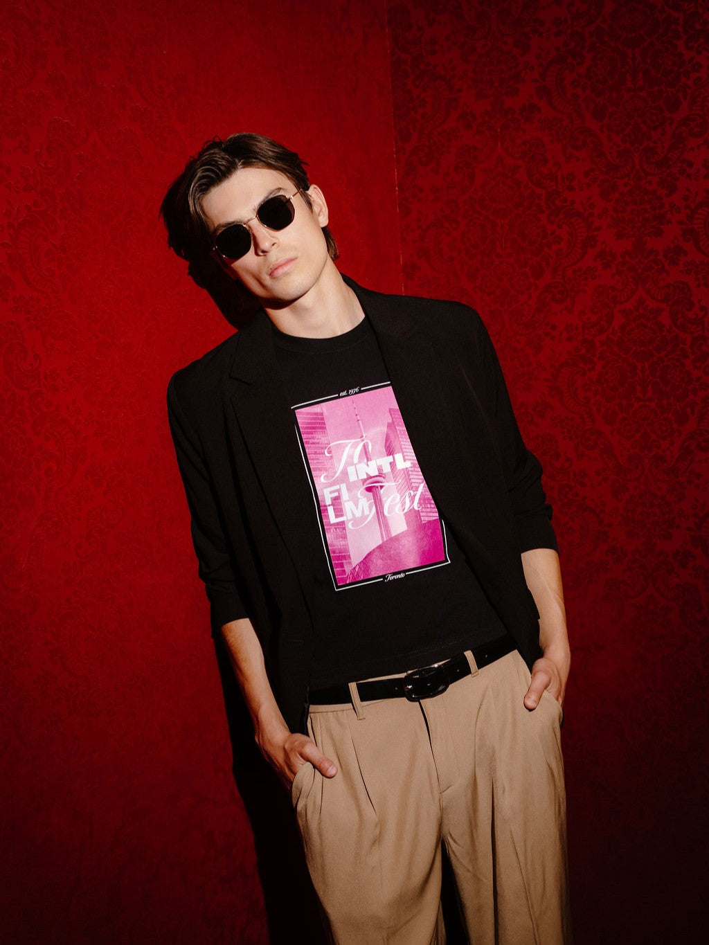 A man wearing a black long-sleeve blazer over a black graphic t-shirt with a pink and white print, paired with beige trousers and sunglasses, posing against a textured red wall.