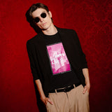 A man wearing a black long-sleeve blazer over a black graphic t-shirt with a pink and white print, paired with beige trousers and sunglasses, posing against a textured red wall.