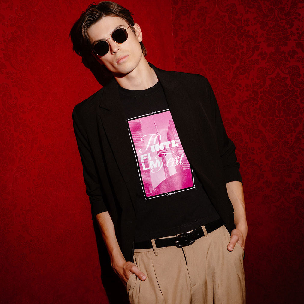 A man wearing a black long-sleeve blazer over a black graphic t-shirt with a pink and white print, paired with beige trousers and sunglasses, posing against a textured red wall.