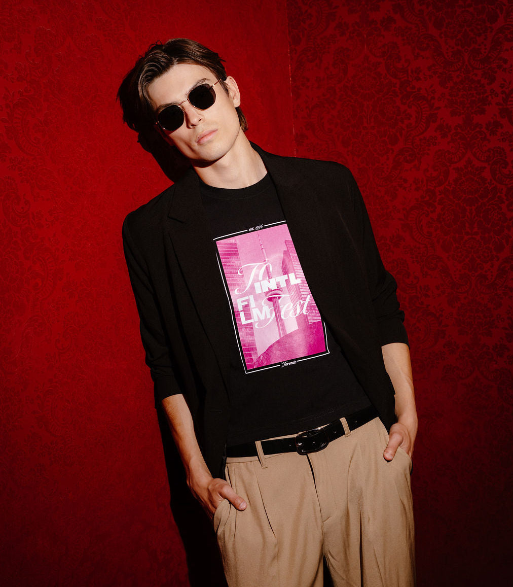 A man wearing a black long-sleeve blazer over a black graphic t-shirt with a pink and white print, paired with beige trousers and sunglasses, posing against a textured red wall.