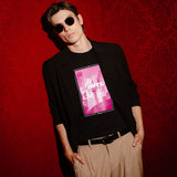 A man wearing a black long-sleeve blazer over a black graphic t-shirt with a pink and white print, paired with beige trousers and sunglasses, posing against a textured red wall.