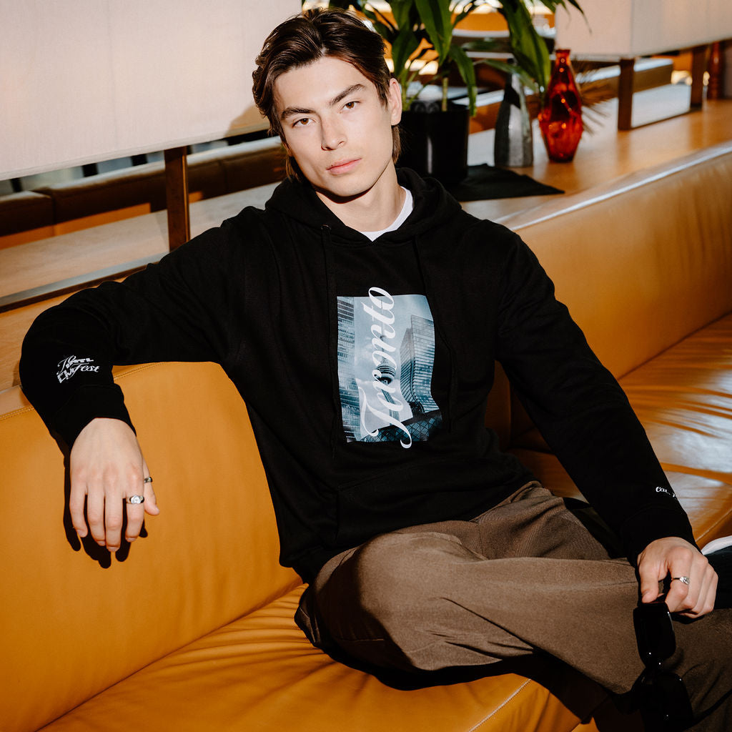 A man wearing a black hoodie with a grey and white graphic print and brown pants, sitting on a tan leather couch in a modern indoor setting.