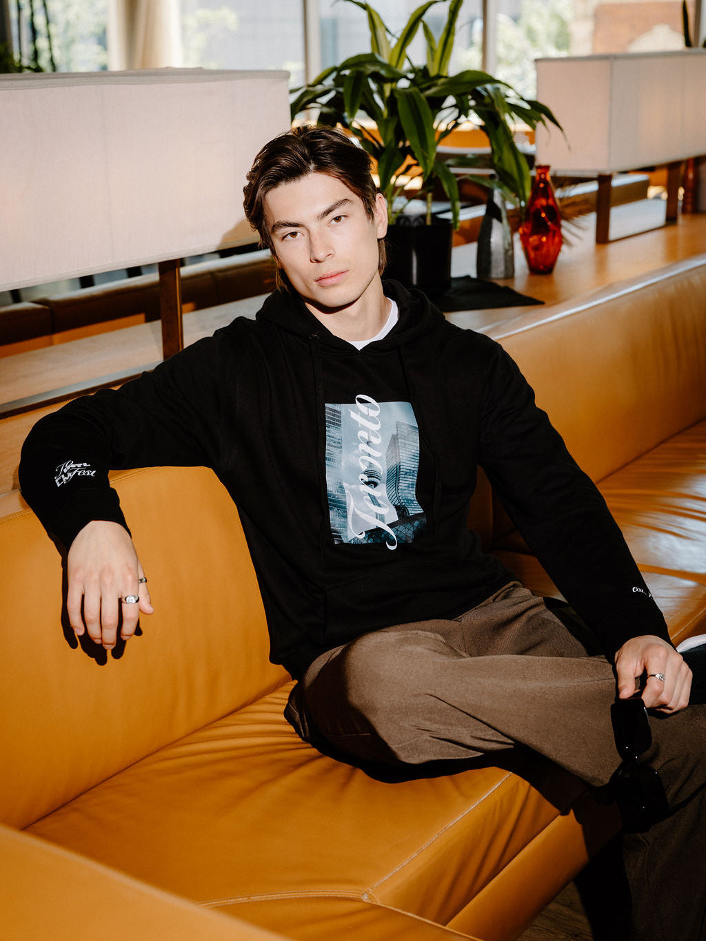 A man wearing a black hoodie with a grey and white graphic print and brown pants, sitting on a tan leather couch in a modern indoor setting.