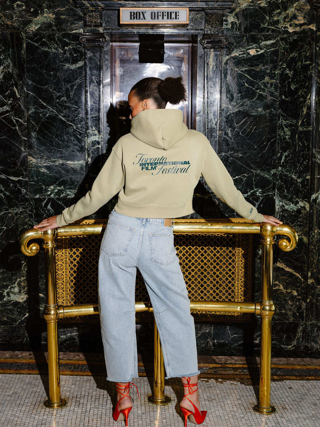 A woman stands with her back to the camera in a lobby, wearing a green hoodie, light-wash jeans, and red high heels, leaning on a gold bench against a dark marble wall.
