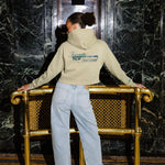 A woman stands with her back to the camera in a lobby, wearing a green hoodie, light-wash jeans, and red high heels, leaning on a gold bench against a dark marble wall.