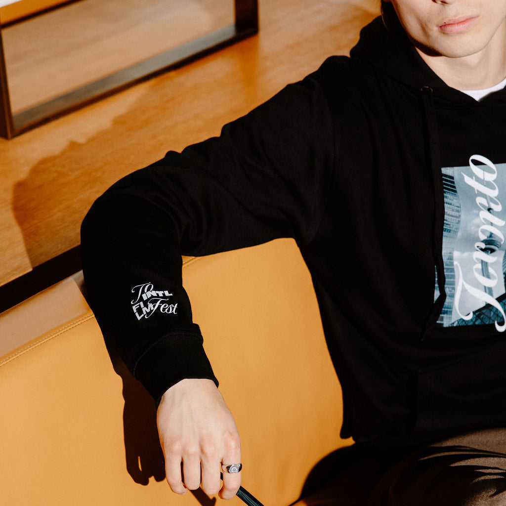 A man wearing a black hoodie with a graphic print and sleeve patch sits on an orange leather couch, holding a pair of black sunglasses.