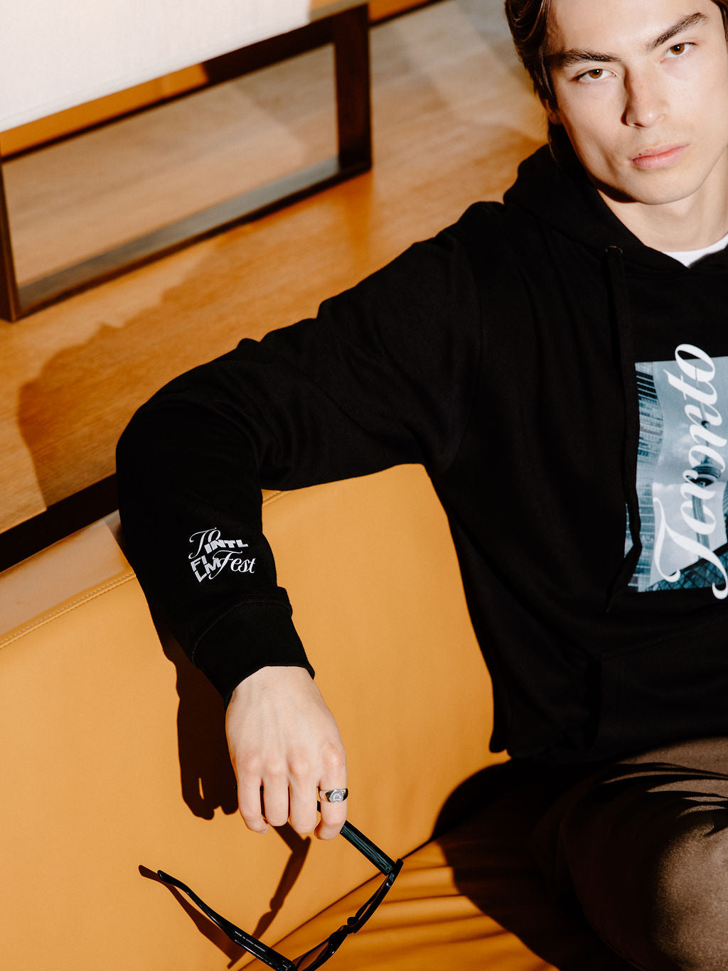 A man wearing a black hoodie with a graphic print and sleeve patch sits on an orange leather couch, holding a pair of black sunglasses.