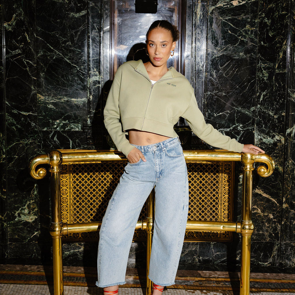 A woman posing in front of a gold-framed bench in a marble hallway, wearing a light sage green cropped zip-up hoodie, light-wash wide-leg jeans, and red high heels.