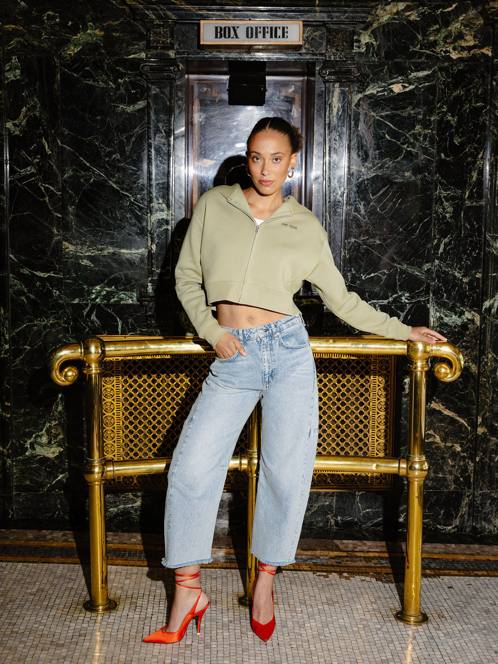 A woman posing in front of a gold-framed bench in a marble hallway, wearing a light sage green cropped zip-up hoodie, light-wash wide-leg jeans, and red high heels.