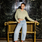 A woman posing in front of a gold-framed bench in a marble hallway, wearing a light sage green cropped zip-up hoodie, light-wash wide-leg jeans, and red high heels.