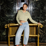 A woman posing in front of a gold-framed bench in a marble hallway, wearing a light sage green cropped zip-up hoodie, light-wash wide-leg jeans, and red high heels.