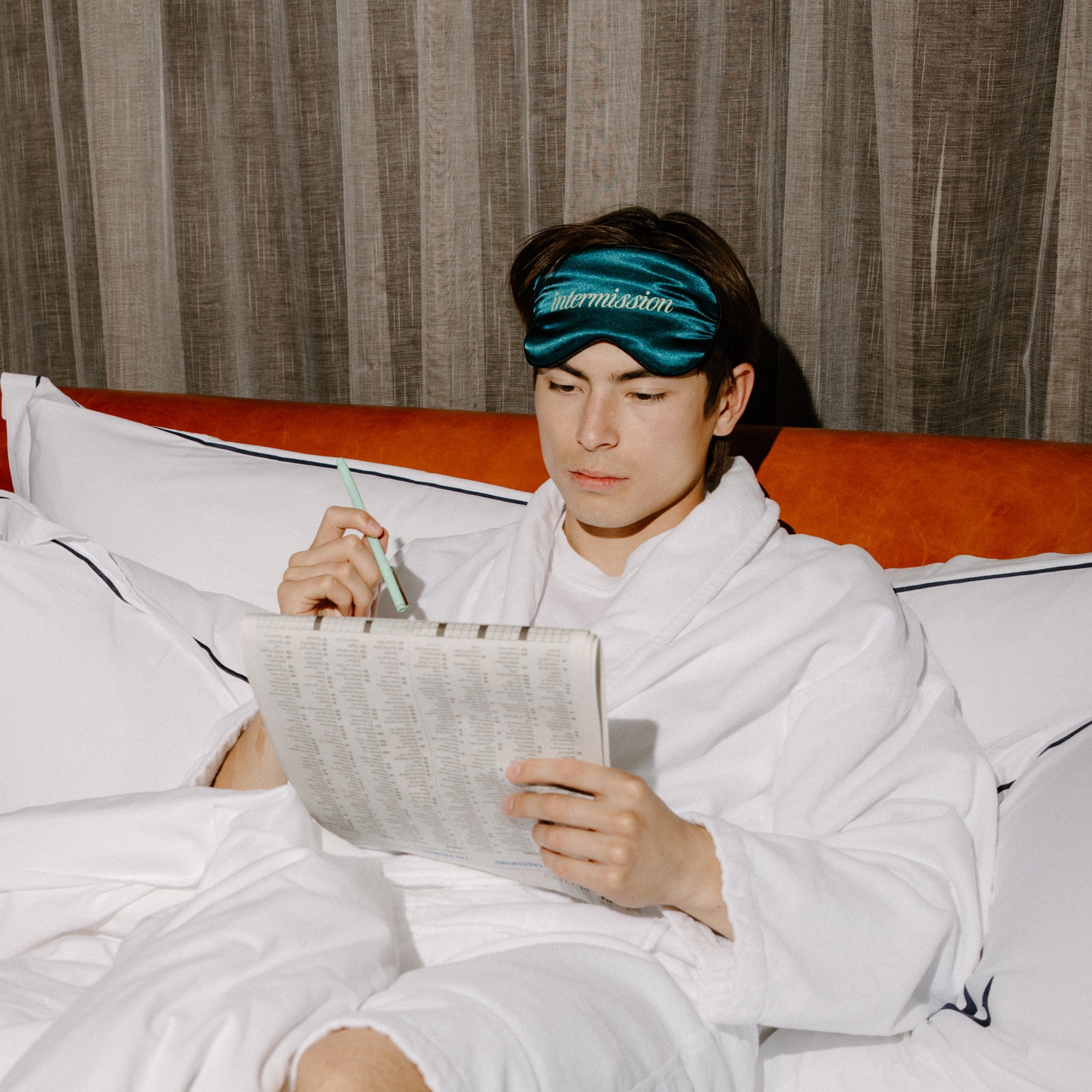A man in a white bathrobe sitting on a bed, wearing a teal sleep mask with the brand name 'intermission' printed in white. He is holding a pen and a sheet of paper, with a textured brown curtain in the background.