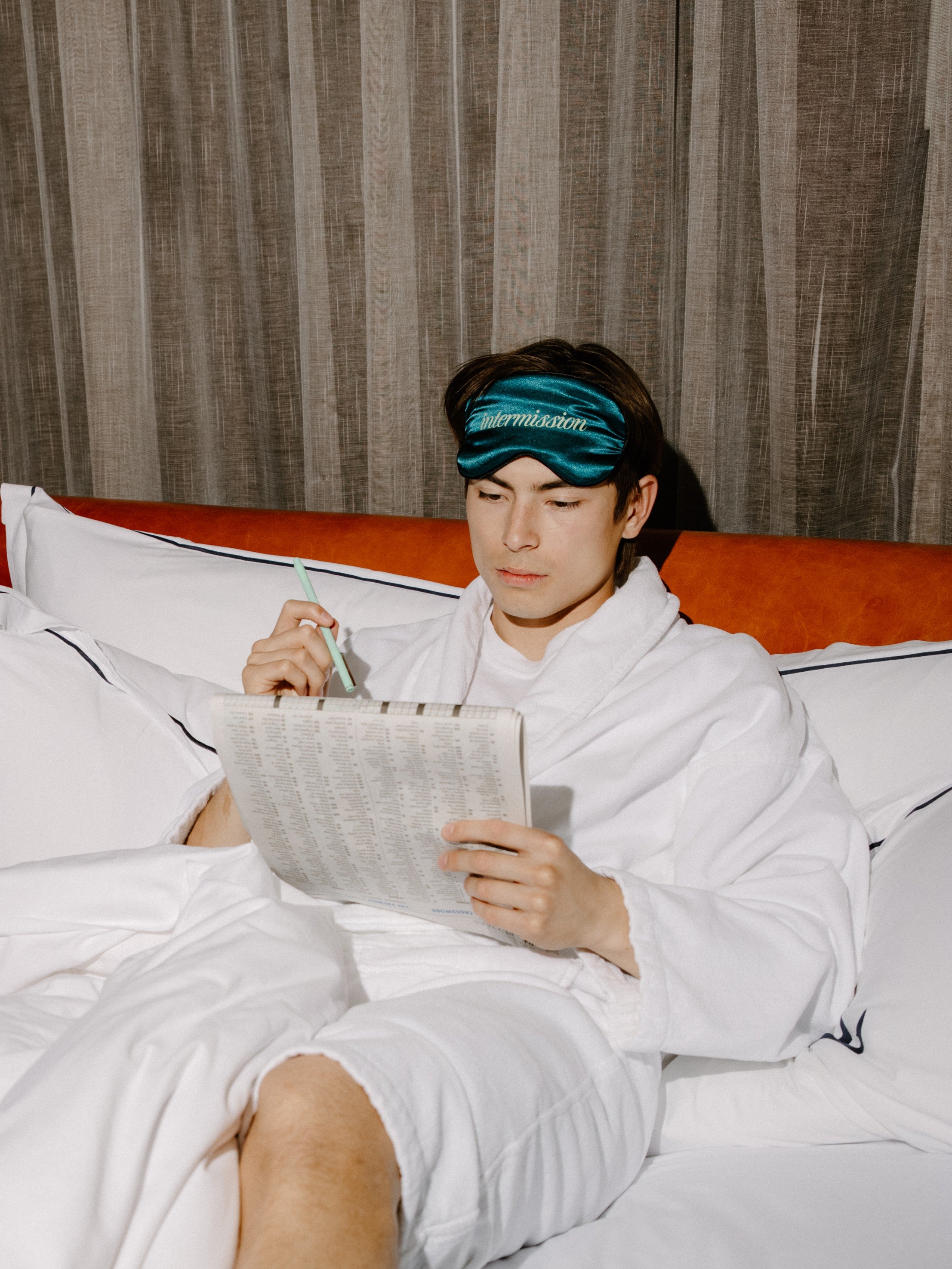 A man in a white bathrobe sitting on a bed, wearing a teal sleep mask with the brand name 'intermission' printed in white. He is holding a pen and a sheet of paper, with a textured brown curtain in the background.