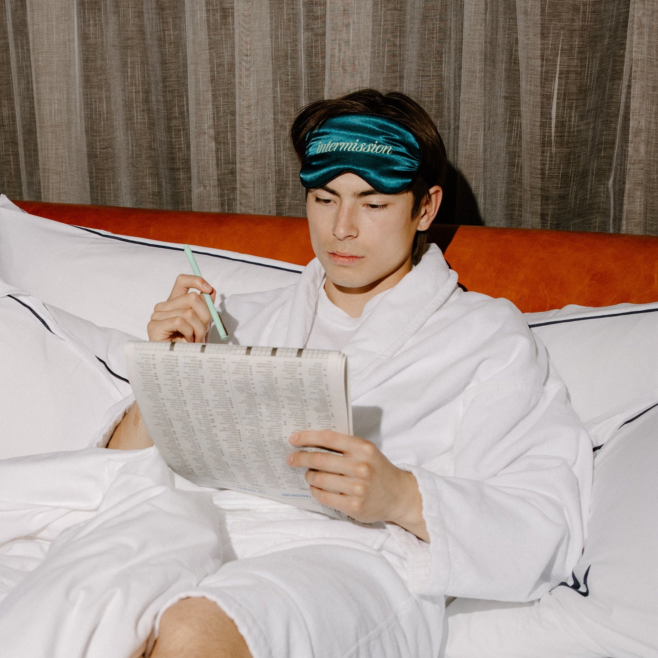 A man in a white bathrobe sitting on a bed, wearing a teal sleep mask with the brand name 'intermission' printed in white. He is holding a pen and a sheet of paper, with a textured brown curtain in the background.