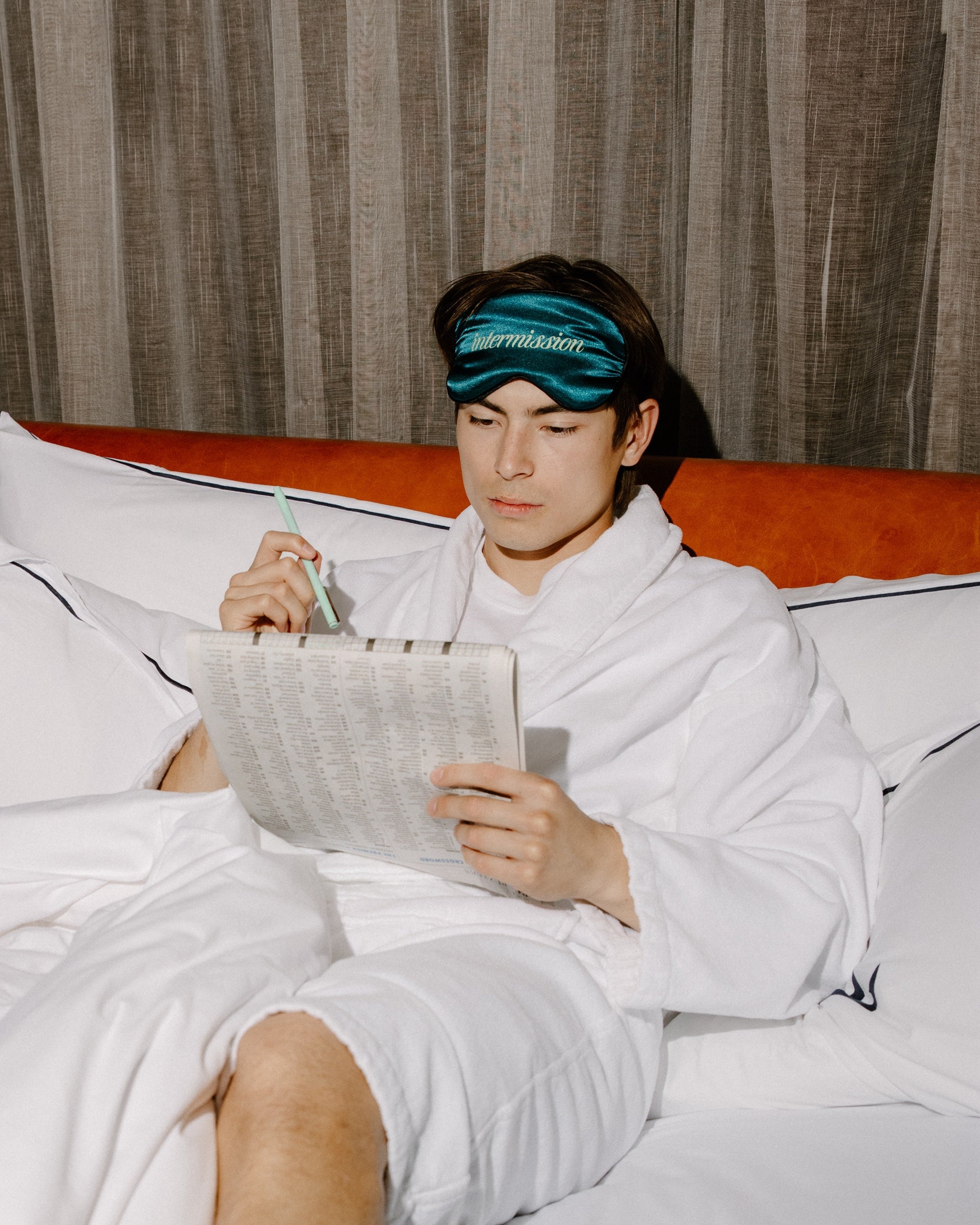 A man in a white bathrobe sitting on a bed, wearing a teal sleep mask with the brand name 'intermission' printed in white. He is holding a pen and a sheet of paper, with a textured brown curtain in the background.