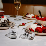 Two clear glass shot glasses with black script text, one standing upright and one lying on its side, placed on a white tablecloth at a dessert table with strawberries, cake, and a wine glass in the background.