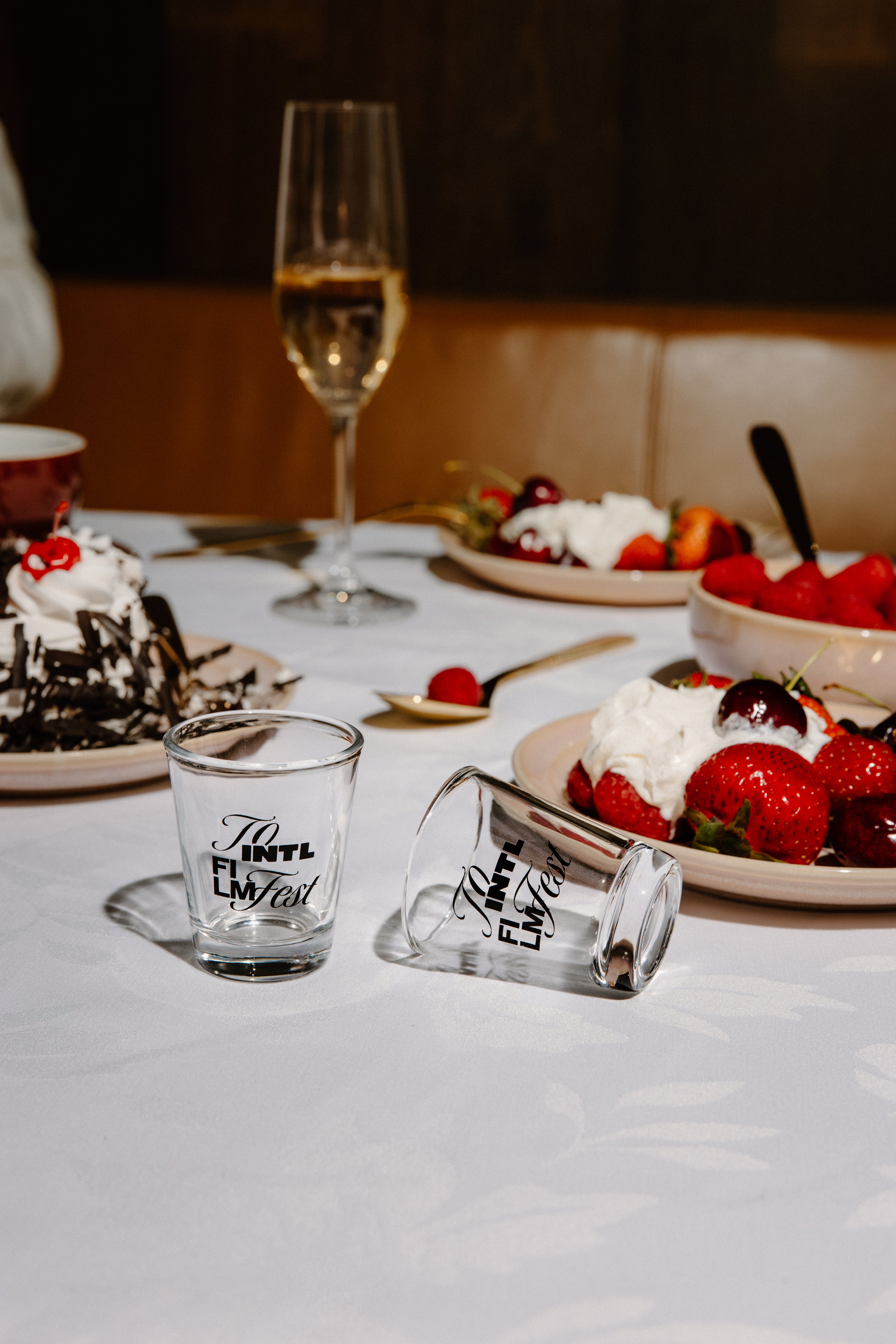 Two clear glass shot glasses with black script text, one standing upright and one lying on its side, placed on a white tablecloth at a dessert table with strawberries, cake, and a wine glass in the background.