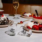 Two clear glass shot glasses with black script text, one standing upright and one lying on its side, placed on a white tablecloth at a dessert table with strawberries, cake, and a wine glass in the background.