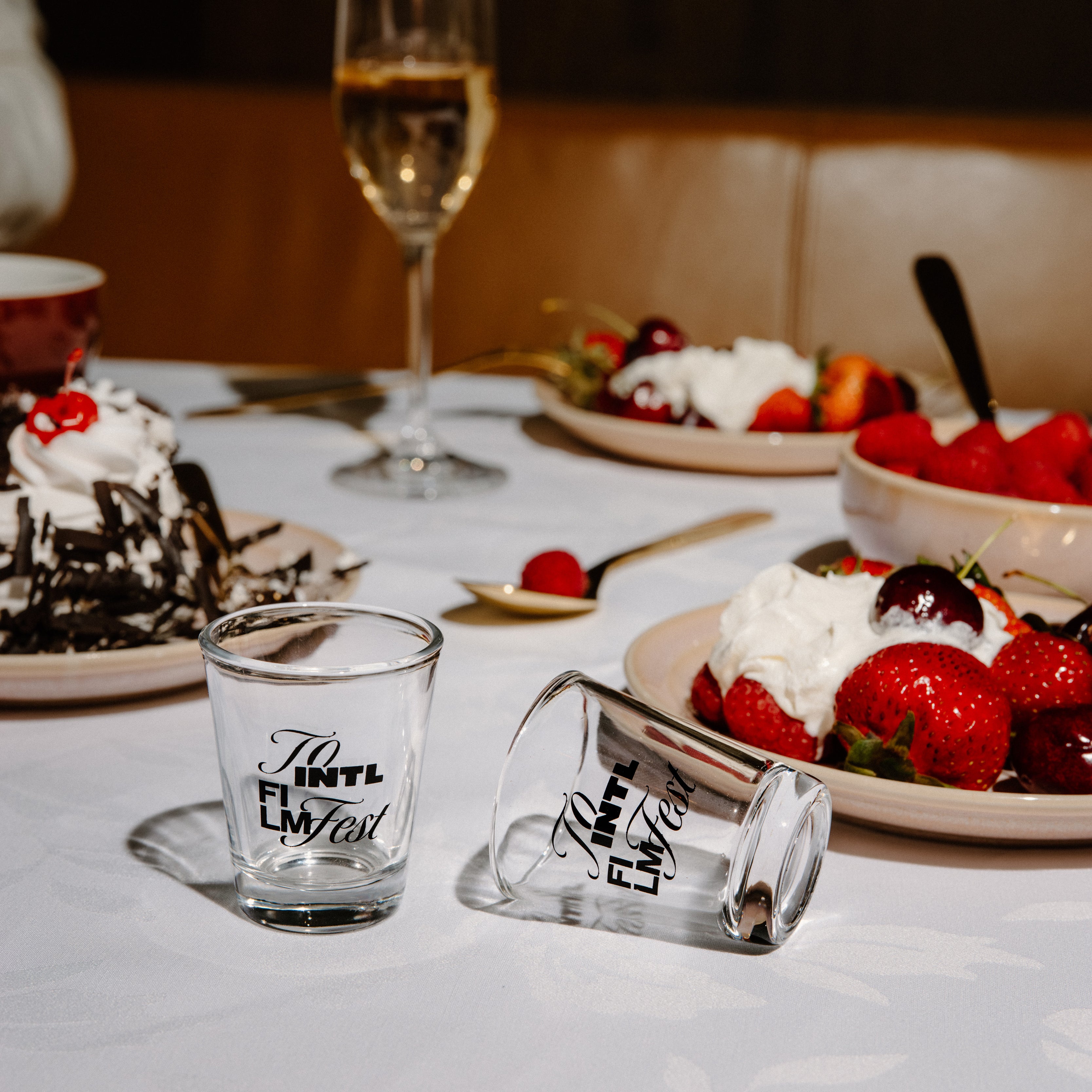 Two clear glass shot glasses with black script text, one standing upright and one lying on its side, placed on a white tablecloth at a dessert table with strawberries, cake, and a wine glass in the background.