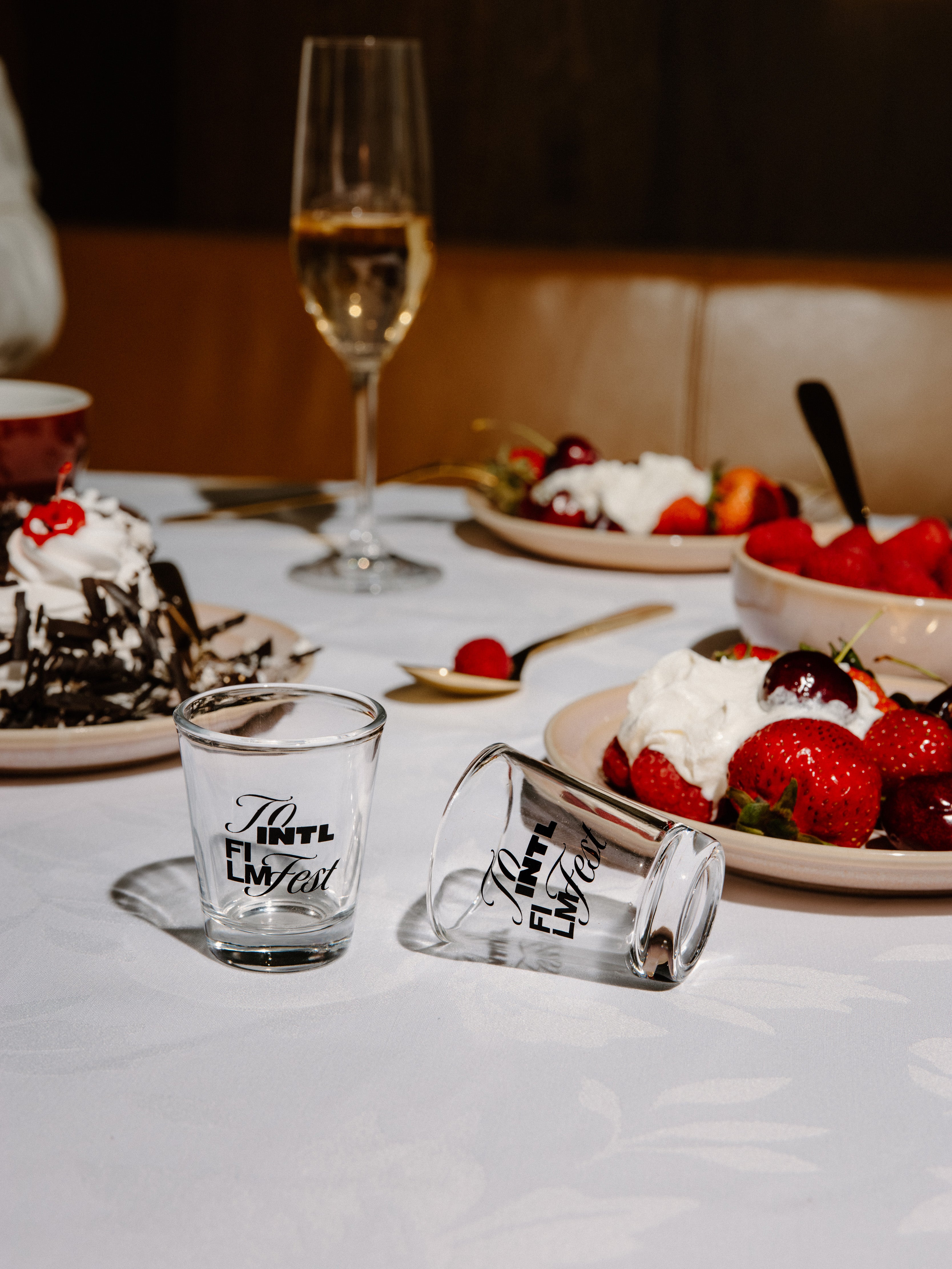 Two clear glass shot glasses with black script text, one standing upright and one lying on its side, placed on a white tablecloth at a dessert table with strawberries, cake, and a wine glass in the background.