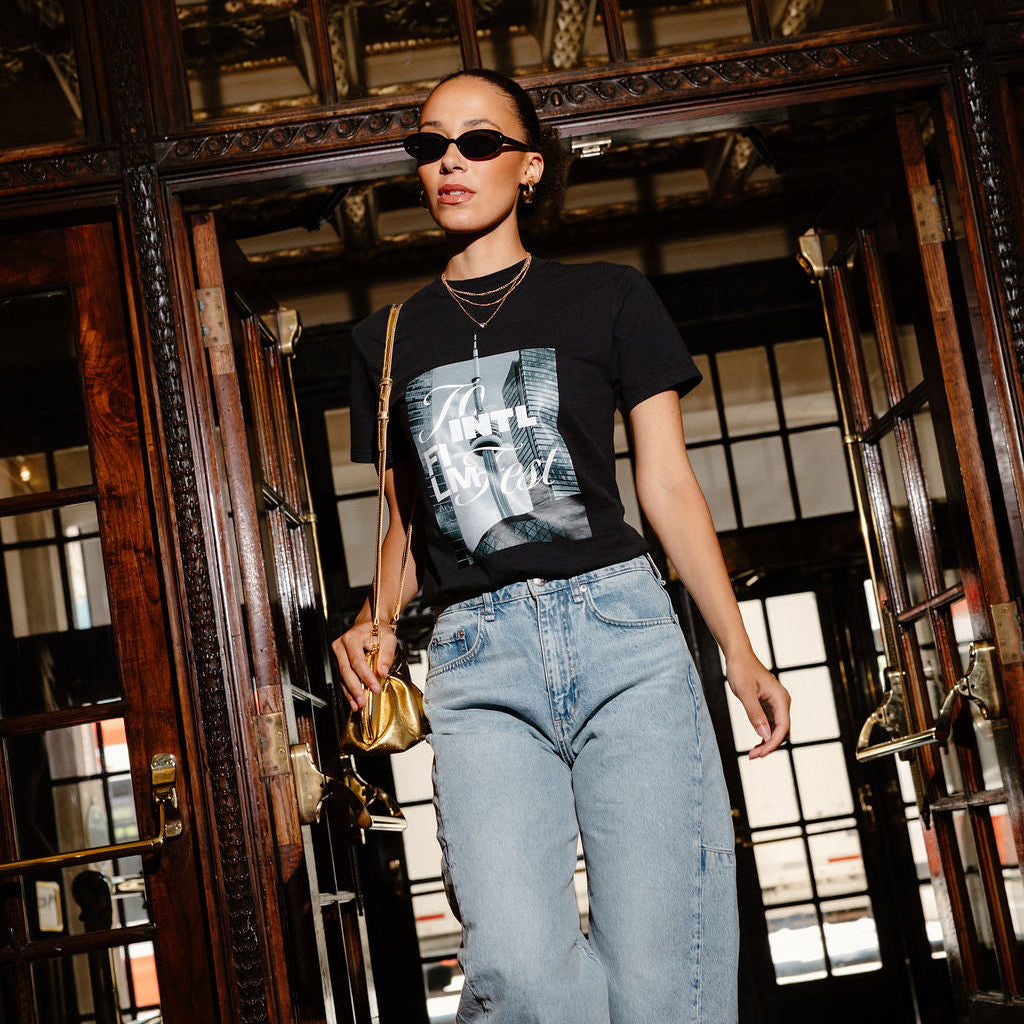 A woman walking through a grand, dark wood doorway wearing a black graphic t-shirt, light-wash blue jeans, sunglasses, and carrying a small gold handbag.