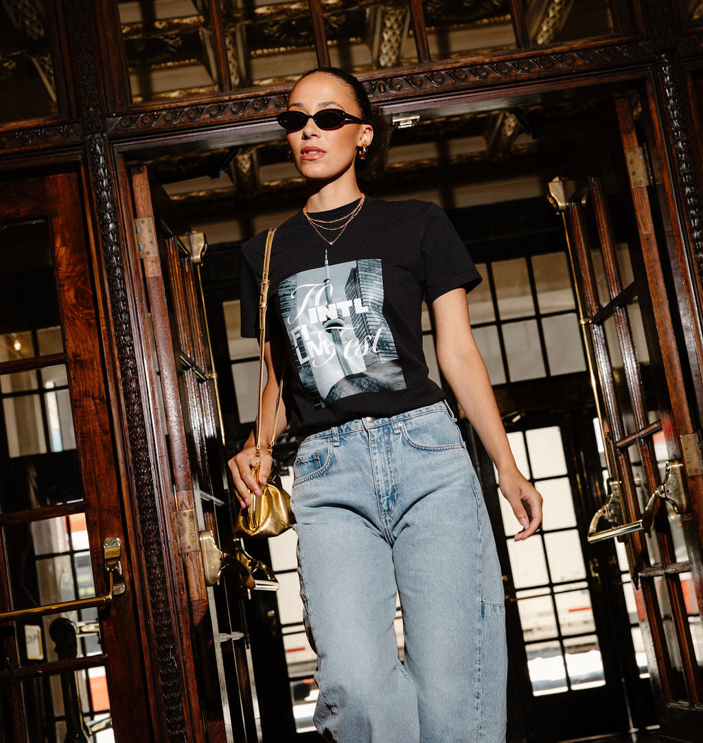 A woman walking through a grand, dark wood doorway wearing a black graphic t-shirt, light-wash blue jeans, sunglasses, and carrying a small gold handbag.